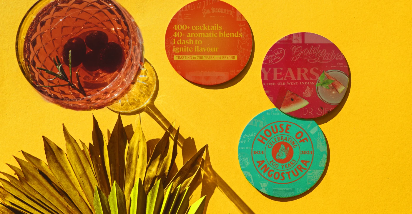 Angostura 200-year coasters in orange, red and teal beside a cocktail on a yellow background.