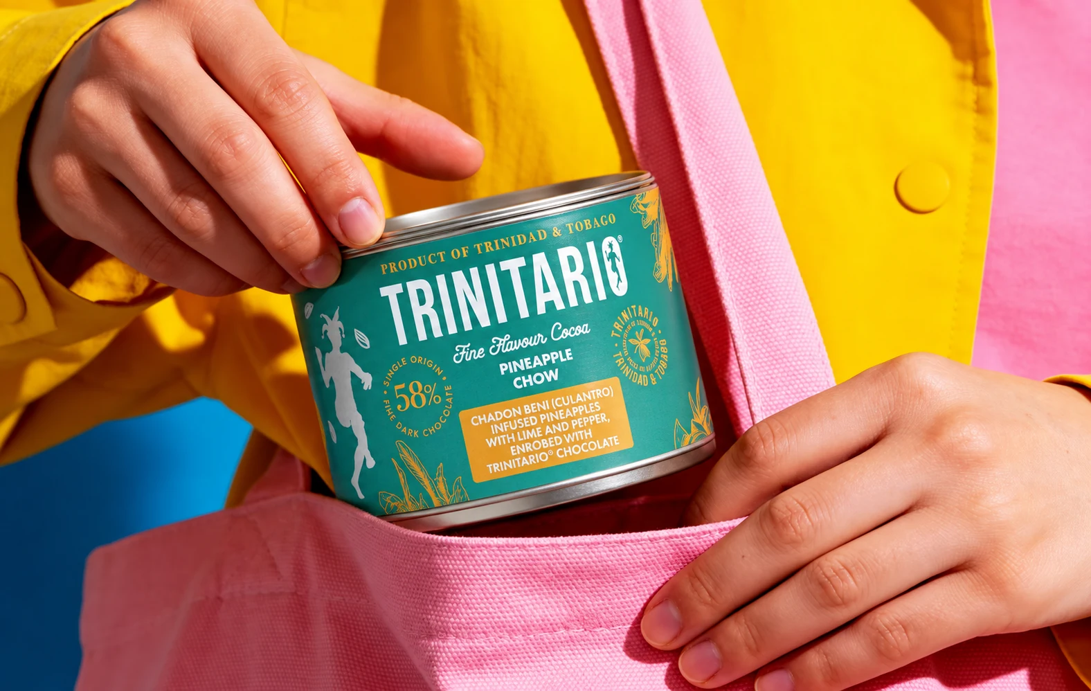 A Trinitario Pineapple Chow tin held above a pink tote bag against a yellow jacket.