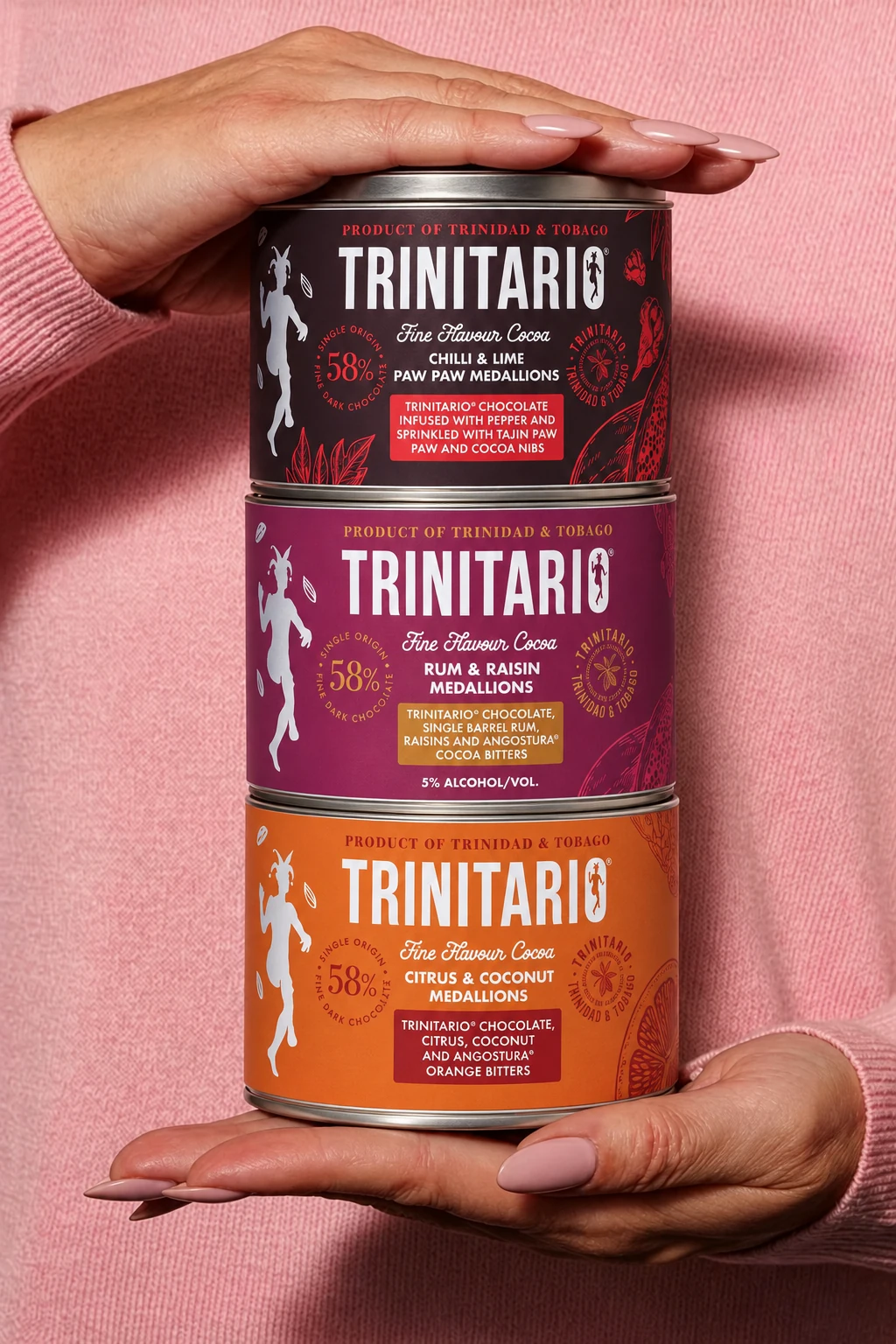 Three Trinitario tins stacked between hands against a pink jumper: Chilli and Lime Paw Paw, Rum and Raisin, and Citrus and Coconut.